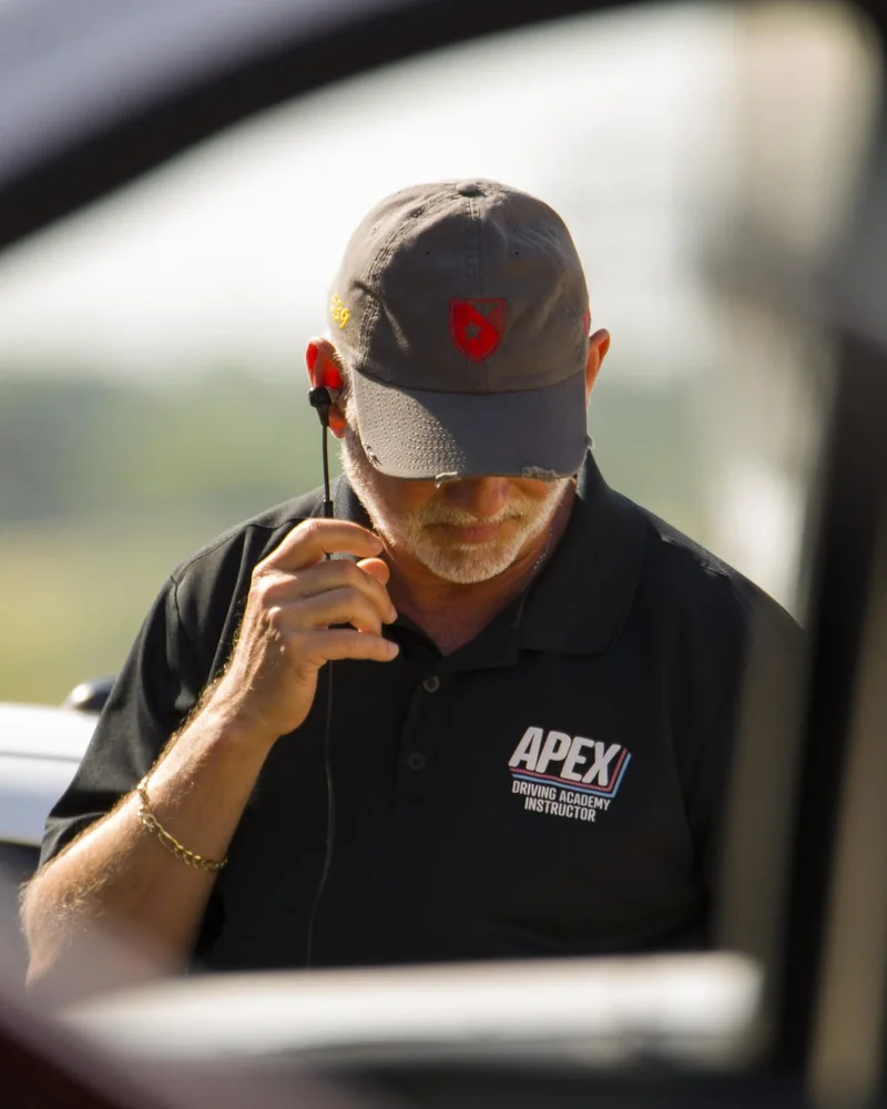 A person in an Apex instructor polo using a handheld radio beside a car
